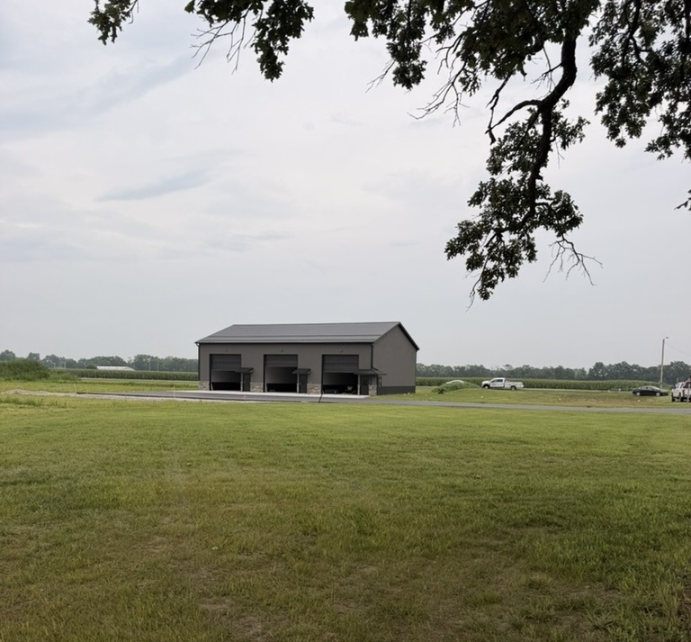 Wide view of Building 1 with fresh asphalt — gray siding, black trim, stone wainscoting, and a massive heritage hickory on the DGC common element