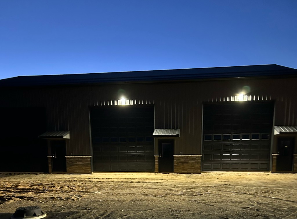 Building 1 at night — wide view of two garage bays with dawn-to-dusk exterior lights on, stone wainscoting, and gravel lot
