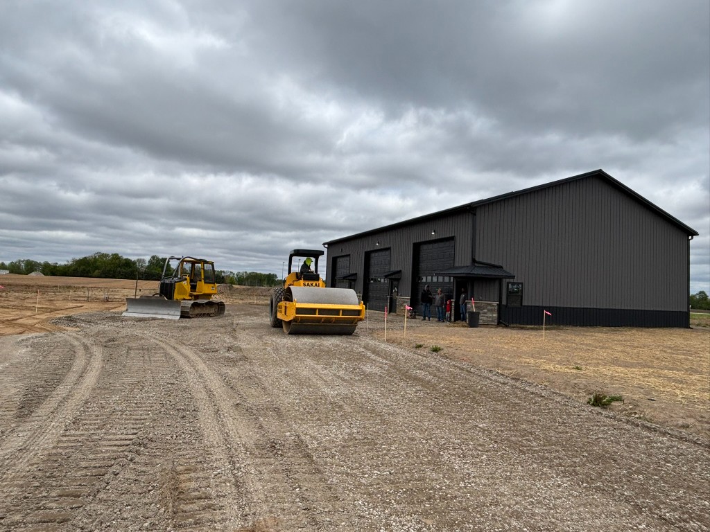 Wide view — Sakai roller and bulldozer compacting the lot with Building 1 and three garage bays behind